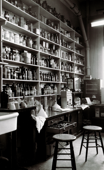 Interior of a pharmacology lab; contains a sink and tabled with drawers underneath, one of which stands open. Stools sit beside this, and along the entire wall are shelves full of pharmaceutical bottles.